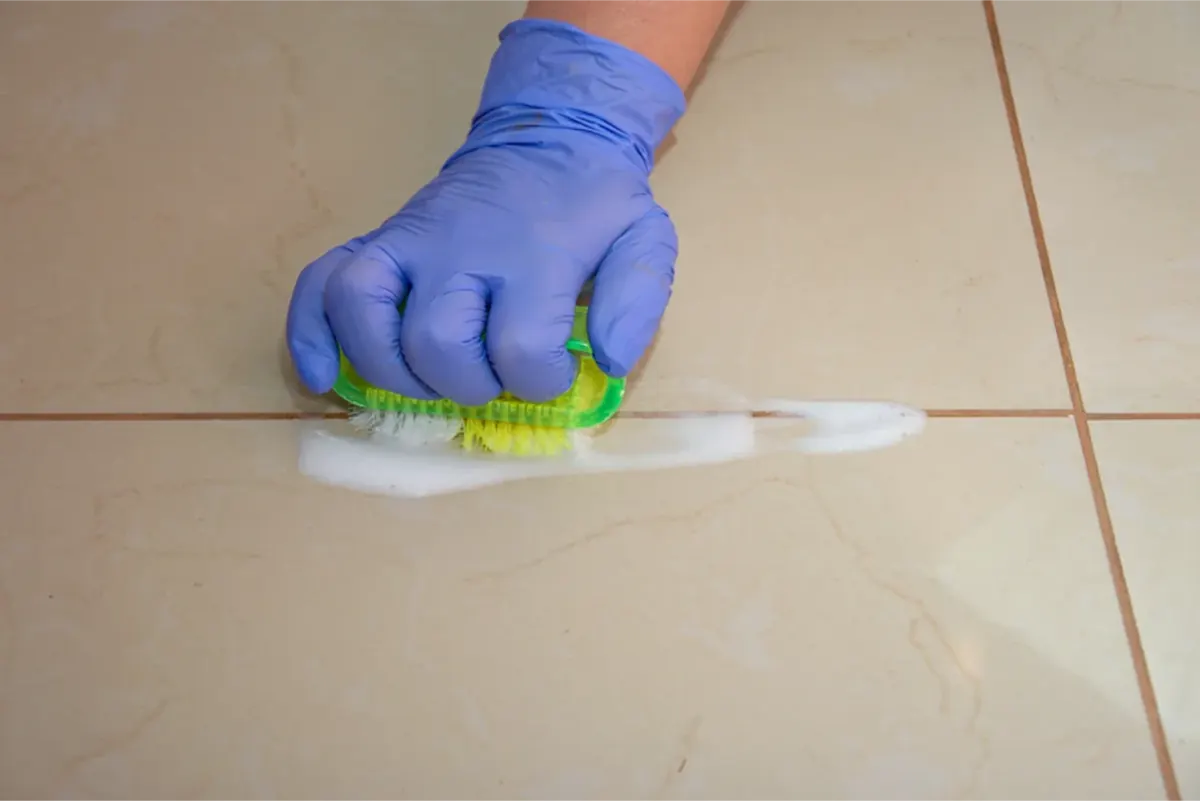 Gloved hand scrubbing tile grout lines with a brush and cleaning foam